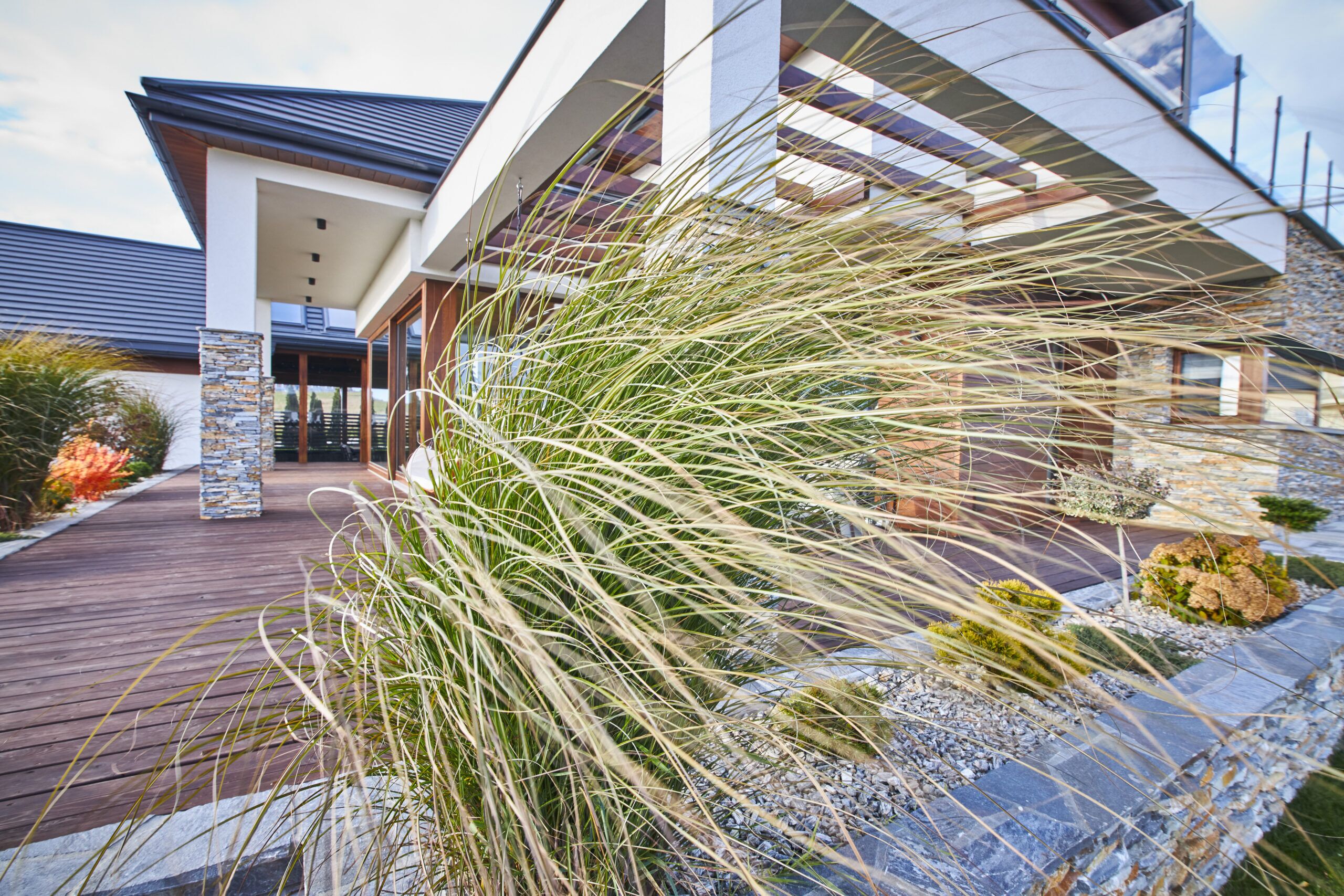 dekoracyjna rama wokół dużego drewnianego tarasu z pergolą i designerska zieleń oraz kamień łupek to pomysł na piękny projekt dużego domu w Nowym Sączu